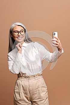 Glad senior woman is using smartphone for video connection isolated on beige, carefree mature gray-haired lady has