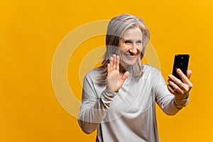 Glad senior woman is using smartphone for video connection isolated on yellow