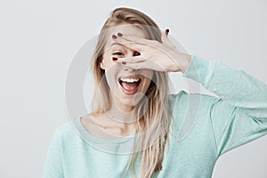 Glad smiling blonde female model hiding her face behind hand, has broad smile, happy to recieve compliments from