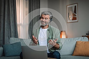 Glad smiling caucasian old man with beard in headphones gesticulates, talks and looks at laptop