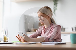 Glad smiling european blond youngsters girl reads message, chatting on smartphone in kitchen interior