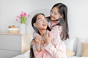 Glad smiling pretty cute asian teenage girl hugging millennial woman in pink pajamas in bedroom interior