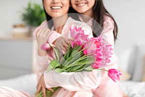 Glad teenage girl hugging millennial japanese lady in pajama giving bouquet of tulips celebrating mother& x27;s day