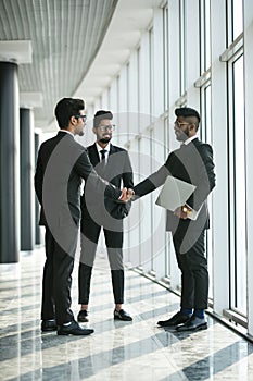 Glad to see you. Two happy young businessmen shaking hands while their colleague smiling in modern office