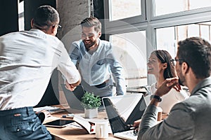 Glad to work with you! Young modern men in smart casual wear shaking hands and smiling while working in the creative office