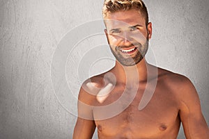 Glad topless male with trendy hairstyle and bristle having strong body builduing posing against grey background with happy express