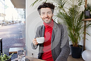 Glad young attractive unshaved brunette guy with trendy haircut holding cup of tea in raised hand and looking positively at camera