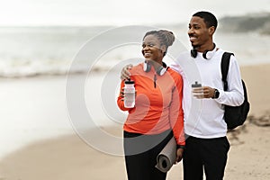 Glad young black couple in sportswear and wireless headphones with bottle water, mats on sea beach
