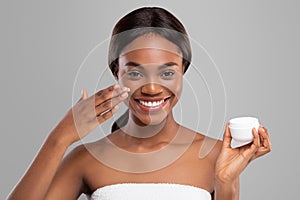 Glad young black woman with perfect skin applying moisturizing anti-aging cream on her face and hold jar