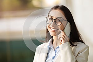 Glad young caucasian businesswoman in beige suit talking on mobile smartphone on office space