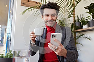 Glad young handsome dark haired bearded guy looking cheerfully on his smartphone and keeping cup in raised hand, sitting over cafe