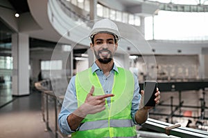 Glad young islamic engineer builder with beard in protective clothing and hard hat points finger at smartphone