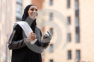 Glad young muslim lady in hijab and sportswear with phone on shoulder with cocktail cup and towel at street