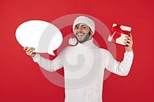 Gladful hipster man wears santa claus hat holding gift box and speech bubble over red background.