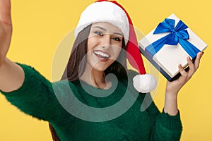 Gladful young brunette woman wear santa claus hat and green pullover take selfie with gift box over yellow background.