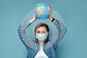 Gladful young woman in medical mask holding world globe over head on blue background.