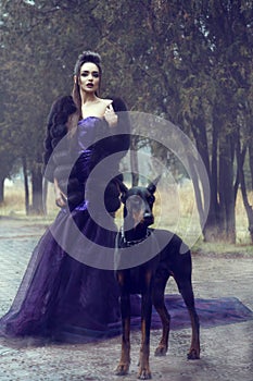 Glam lady in luxurious sequin violet evening gown and fur coat standing on the alley in the park with her Doberman pinscher dog