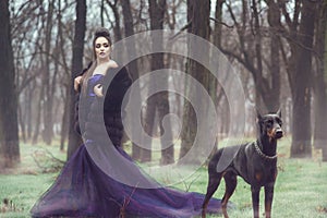 Glam lady in luxurious sequin violet evening gown and fur coat standing in the woods with her Doberman pinscher dog