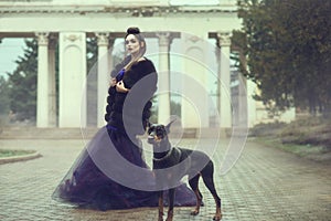 Glam lady wearing luxurious sequin violet evening gown and fur coat standing on the alley in the park with her Doberman pinscher