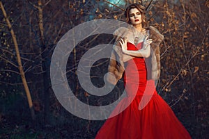 Glam model with updo hair and beautiful make up wearing posh red fishtail dress and luxurious mink vest standing in dry bushes