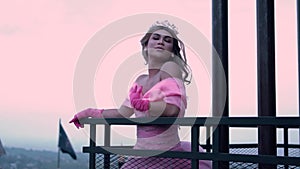 Glamor princess standing on the rooftop in front of the black fence while wearing a crown and a pink dress