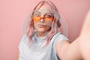 Glamorous 30s woman blogger in bright orange stylish glasses looks at the camera and takes a selfie isolated over pink