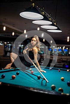 Glamorous brunette woman holding a cue and sitting on the billiard table