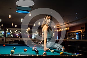 Glamorous brunette woman holding a cue and sitting on the billiard table