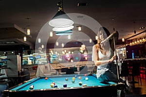 Glamorous brunette woman holding a cue and sitting on the billiard table