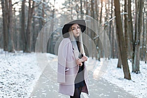 Glamorous cute beautiful young woman in stylish warm glamorous clothes in an elegant hat is stands on the road in a snowy park