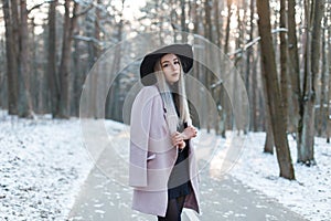 Glamorous cute beautiful young woman in stylish warm glamorous clothes in an elegant hat is stands on the road in a snowy park on