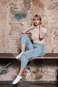 Glamorous fashion model of a pretty young blonde woman with beautiful lips in stylish blue jeans in a trendy t-shirt rests near a