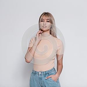 Glamorous fashion model of a young blonde woman in stylish blue jeans in a trendy shirt with a hairstyle stands near a white wall