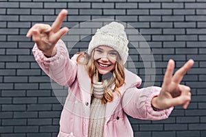 Glamorous female model in pink winter jacket posing with peace sign. Outdoor shot of laughing blithesome blonde woman in