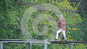 Glamorous handsome blond man dances and sings standing on suspension bridge over river in beautiful pictorial place