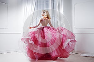 Glamorous lady in a chic pink dress with a train. Studio portrait in white interior, back view