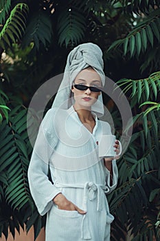 Glamorous lifestyle. Woman in a dressing gown with a white towel on her head and elegant sunglasses posing with a cup of coffee