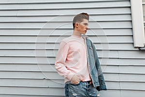 Glamorous pretty young man model in trendy pink sweatshirt in fashion denim blue youth clothes posing near white wall outdoors.