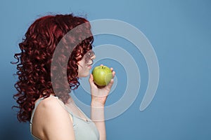 Glamorous woman with red curly hair in grey top standing sideways and holding green apple on blue studio wall background