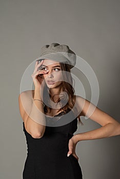 Glamorous young woman wears fashionable dress posing with fluttering hair at grey wall. Girl put forward hand