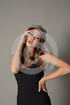 Glamorous young woman wears fashionable dress posing with fluttering hair at grey wall. Girl put forward hand