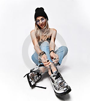 Glamour blonde girl in knitted hat, sunglasses and jeans is sitting on the floor tying shoelaces of her brutal boots