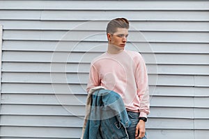 Glamour fashionable young man in pink stylish sweatshirt with denim blue jacket in jeans stands near white vintage building on