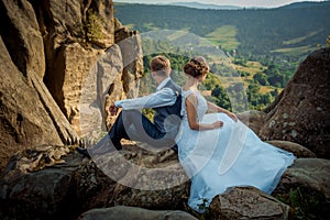 Glamour newlywed couple is sitting on the rock back-to-back and enjoying the landscape view during the sunny day.