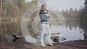 Glamour woman in white pants, jeans jacket and sunglasses standing on the riverbank. The girl is cold, she rubbing her