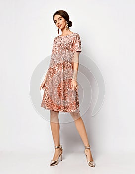 Glamour young female with stylish hairdo and thick earrings demonstrating sparkly short coral pink dress and silver shoes
