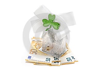 Glamourous home with glitter and white bow with green shamrock on stack of cash on white background. Buying high value house