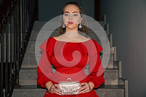 glamourous young beautiful woman in red dress, with silver bag and silver earrings is sitting on the staircase photo