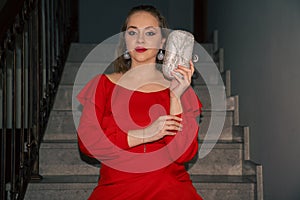 glamourous young beautiful woman in red dress, with silver bag and silver earrings is sitting on the staircase photo