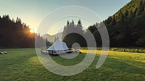 Glamping tent in green meadow surrounded by fir tree forest at sunrise